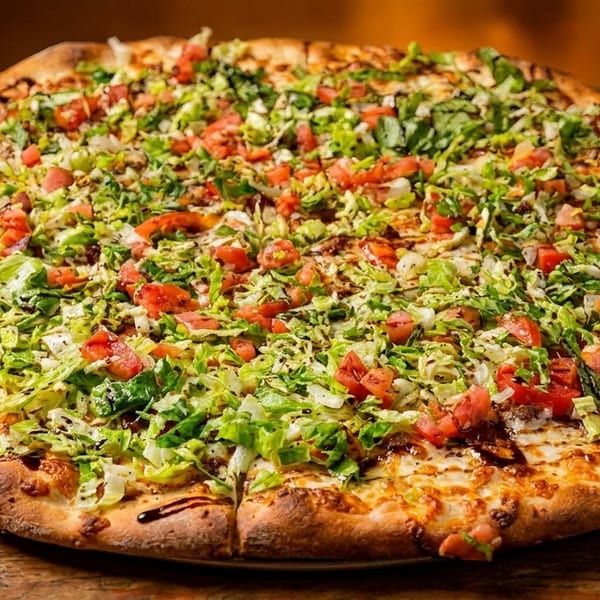A large pizza topped with shredded lettuce, diced tomatoes, and a balsamic glaze on a wooden table.