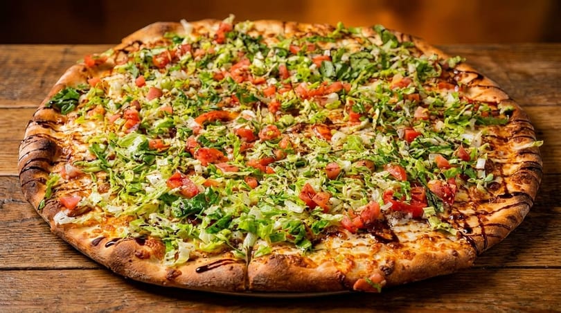 A large pizza topped with shredded lettuce, diced tomatoes, and a balsamic glaze on a wooden table.