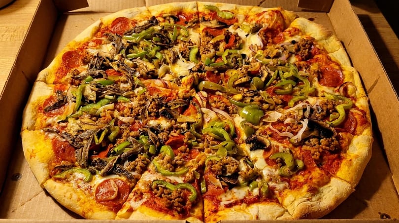 A whole pizza in a cardboard box, topped with pepperoni, sausage, green peppers, onions, and mushrooms.