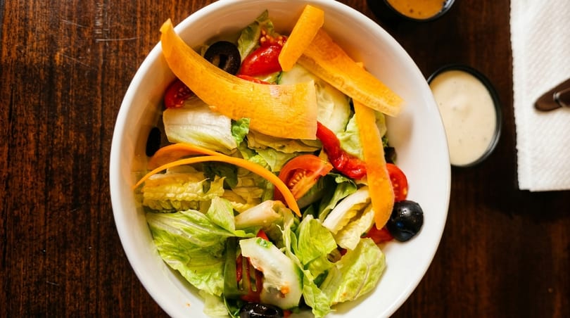 Overhead view of a fresh garden salad with lettuce, tomatoes, olives, and carrot strips in a white bowl.
