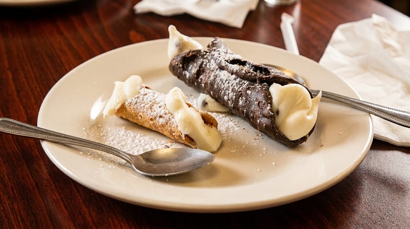 Two cannoli, one chocolate and one classic, dusted with powdered sugar on a white plate.