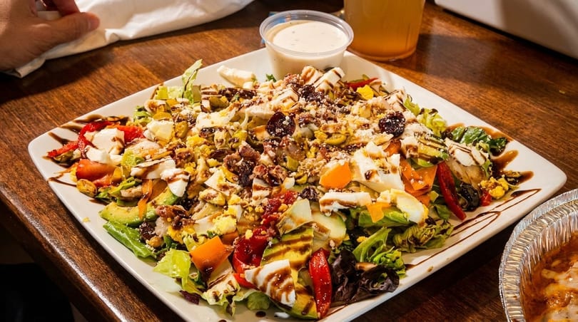 A large, colorful salad with mixed greens, avocado, cheese, eggs, and balsamic glaze.